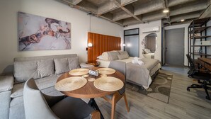 A modern, well-lit room with a sofa, a bed, a dining table with placemats, and a large abstract painting on the wall. - Epic Suites x Ahead (Pereira)