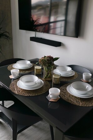 A dining table set with white plates, bowls, and cups, accompanied by a vase of flowers and candles. - Epic Suites x Ahead (Pereira)
