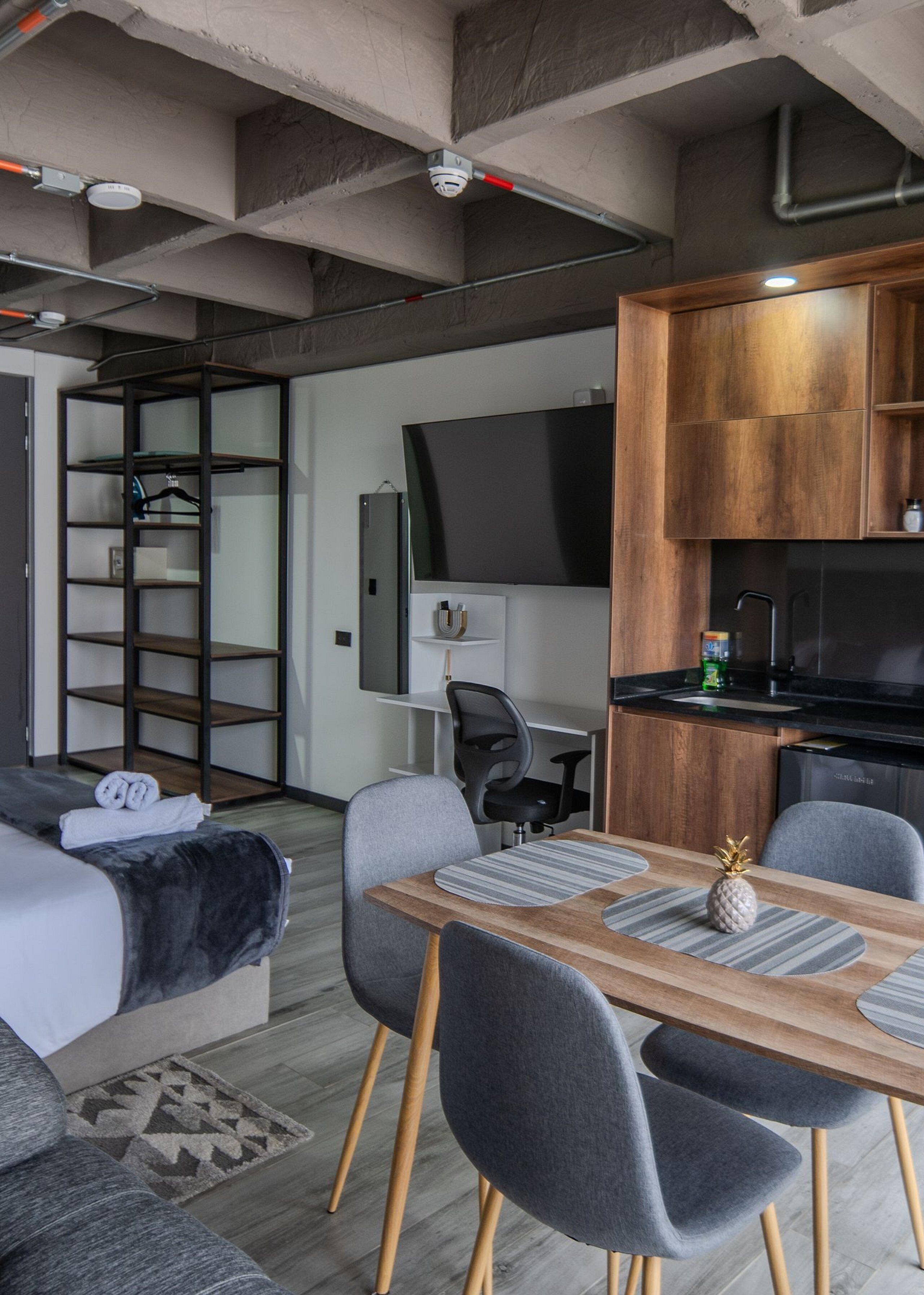 A modern hotel room with a sofa bed, a dining table with chairs, a kitchen area with a TV, and a wooden cabinet.