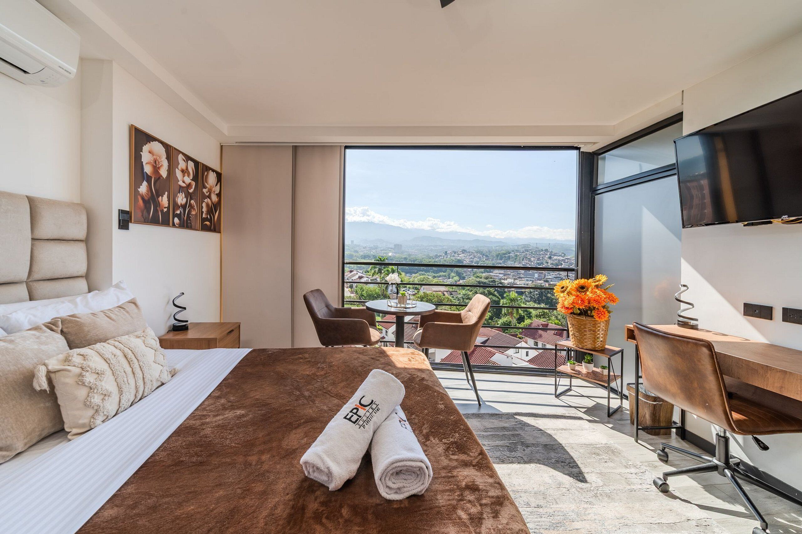 Ein Hotelzimmer mit Bett, Schreibtisch, Sessel, Fernseher und einem Balkon mit Blick auf Stadt und Berge.