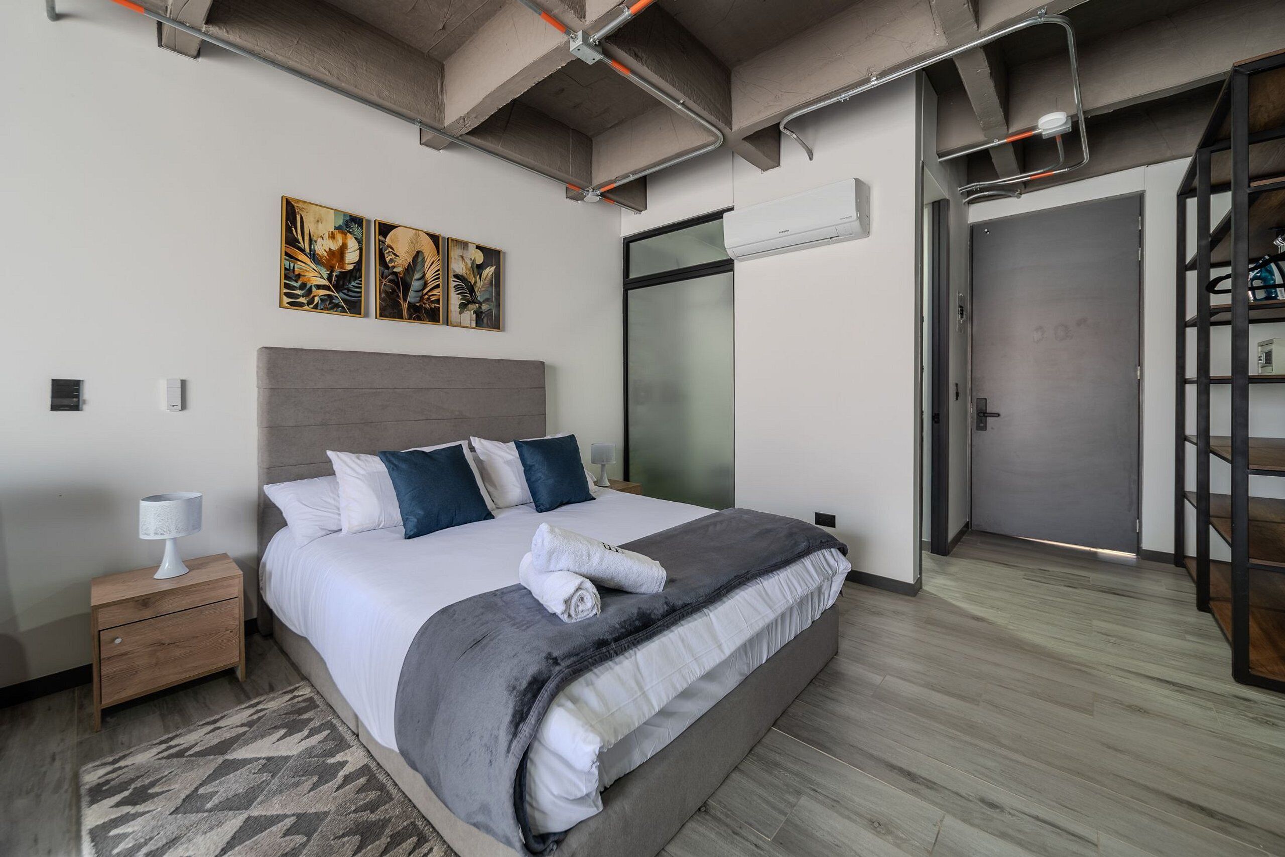Ein modernes Hotelzimmer mit einem großen Bett, einem Nachttisch, einer Wand-Klimaanlage und einer Tür zu einem Flur.