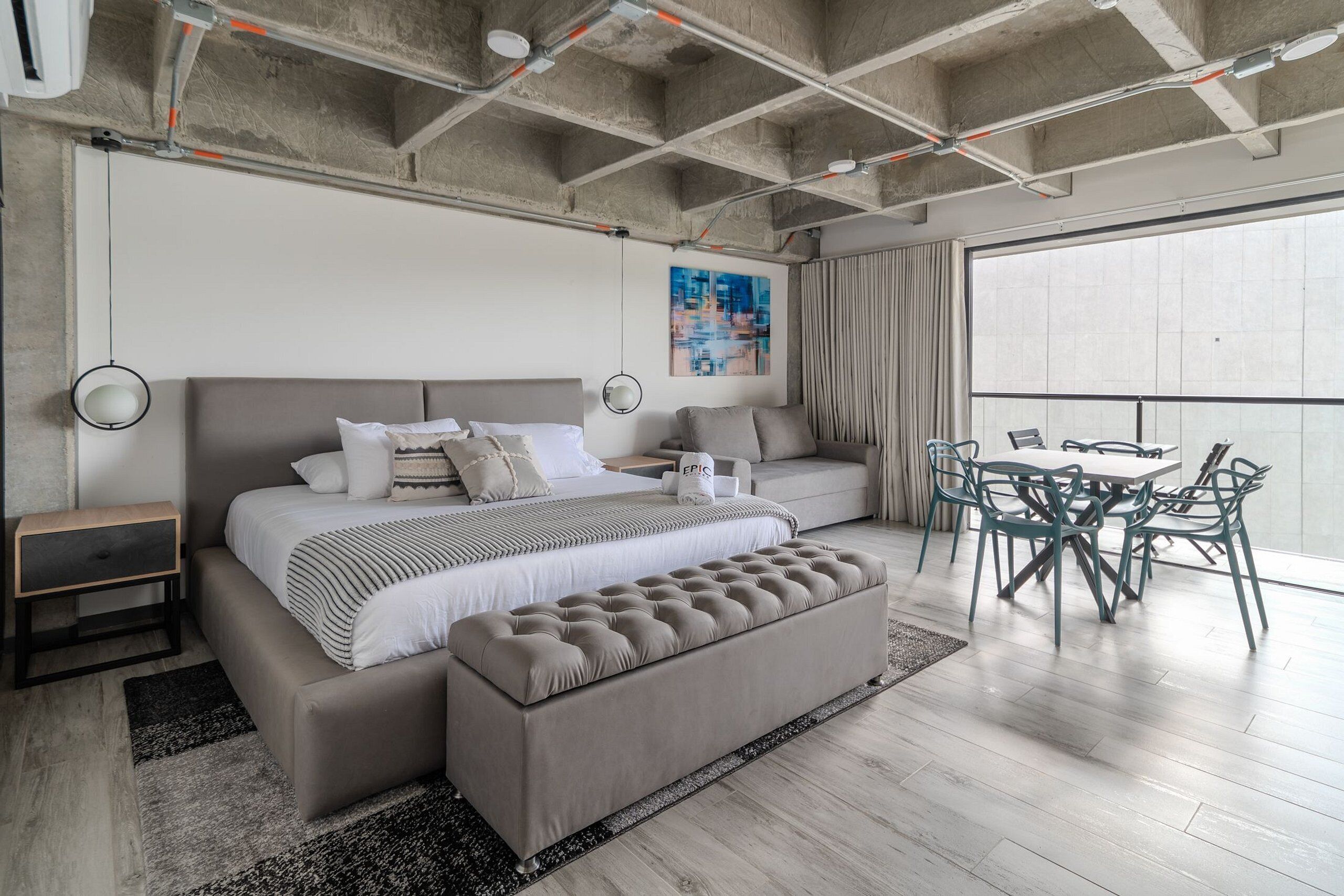 Ein modernes Schlafzimmer mit einem großen Bett, einer Bank, einem Esstisch mit Stühlen und einem großen Fenster.