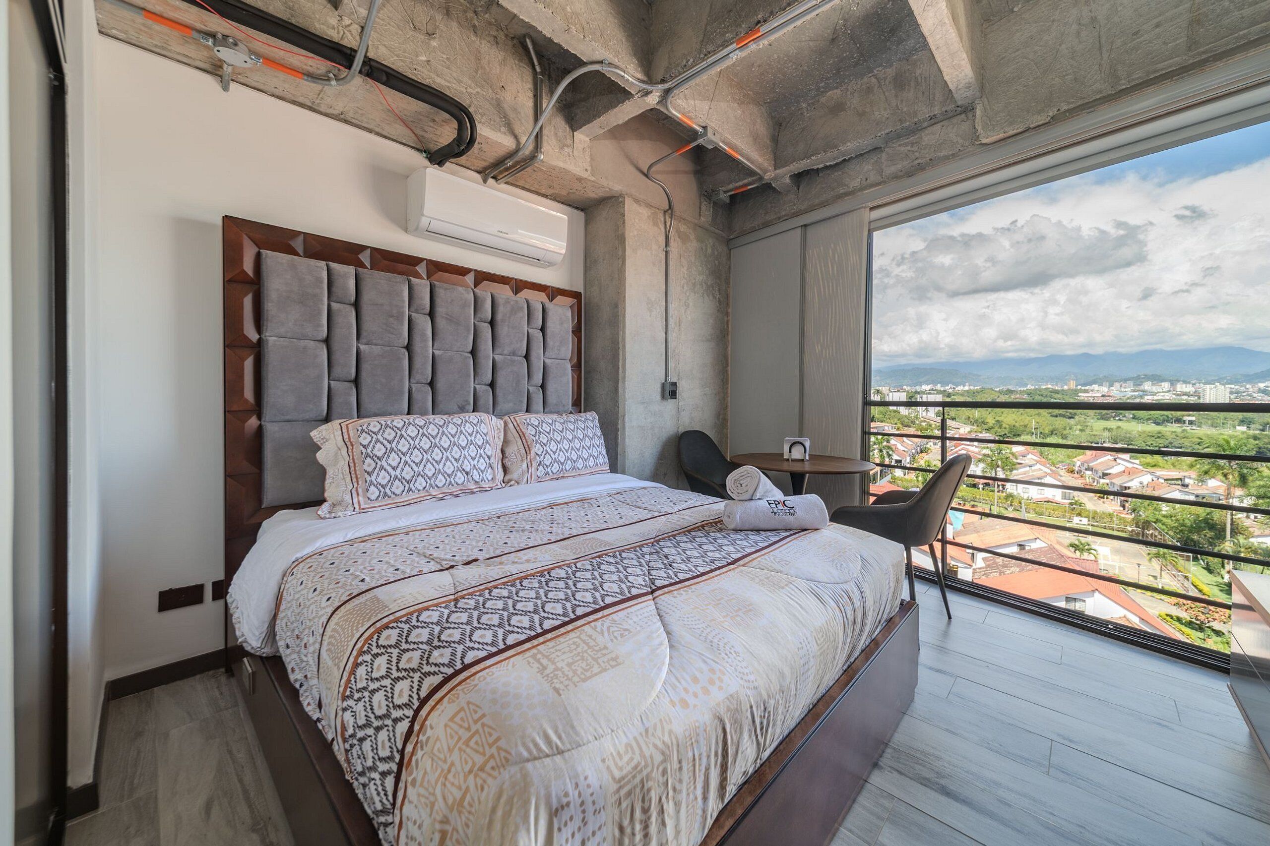 Ein modernes Schlafzimmer mit einem großen Bett, einer Kopfteil mit geometrischen Mustern, Blick auf die Stadt durch einen Balkon und einer Klimaanlage an der Wand.