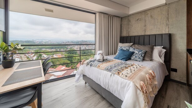 A modern bedroom with a large bed, a desk, and a balcony with a city view. - Epic Suites x Ahead (Pereira)