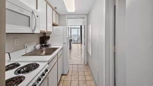 A narrow kitchen with white cabinets, a stove, a microwave, and a sink. A hallway leads to a balcony with a view. - Atlantica 903, Oceanfront Stay, Myrtle Beach Rental (Myrtle Beach)