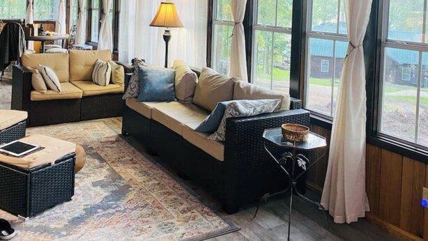A cozy living room with a sofa, a small table, and a lamp. There are large windows with curtains, and a ceiling fan is visible. - Rustic Cozy cabin with water and dock access  (Florien)