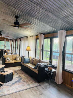 Ein gemütliches Wohnzimmer mit einer Couch, einem kleinen Tisch und einer Lampe. Große Fenster mit Vorhängen sind zu sehen, außerdem ein Deckenventilator.