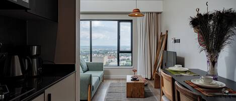 A modern kitchen with a coffee maker, a dining area with a table set for two, a sofa, and a view of the city through large windows.