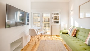 A modern living room with a green sofa, a flat-screen TV mounted on the wall, a dining table with chairs, and a large window offering a view of the cityscape. - GuestReady - A pinch of heaven in Lisbon (Lisbon)