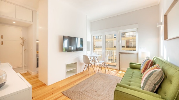 A modern living room with a green sofa, a flat-screen TV, a dining area with chairs, and a view of a building through large windows. - GuestReady - A pinch of heaven in Lisbon (Lisbon)