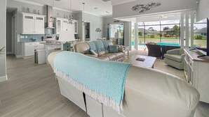 Un ampio soggiorno con un grande divano a forma di L, un tavolino e una TV. C'è un angolo cottura con pensili bianchi e una zona pranzo con tavolo e sedie.