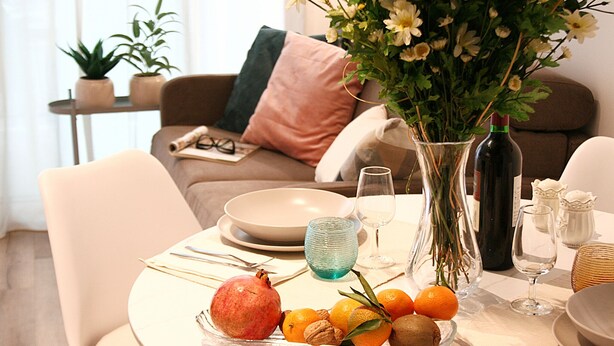 Una mesa de comedor dispuesta con platos, vasos, un jarrón con flores y un frutero.