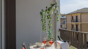 Un balcón con una mesa dispuesta para dos personas, con mantel blanco, dos copas de bebida roja, un cuenco de aperitivos, una planta en maceta y una botella de champán.