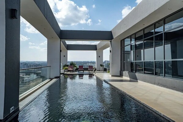 A rooftop infinity pool with a glass balustrade, red lounge chairs, and a view of the cityscape.