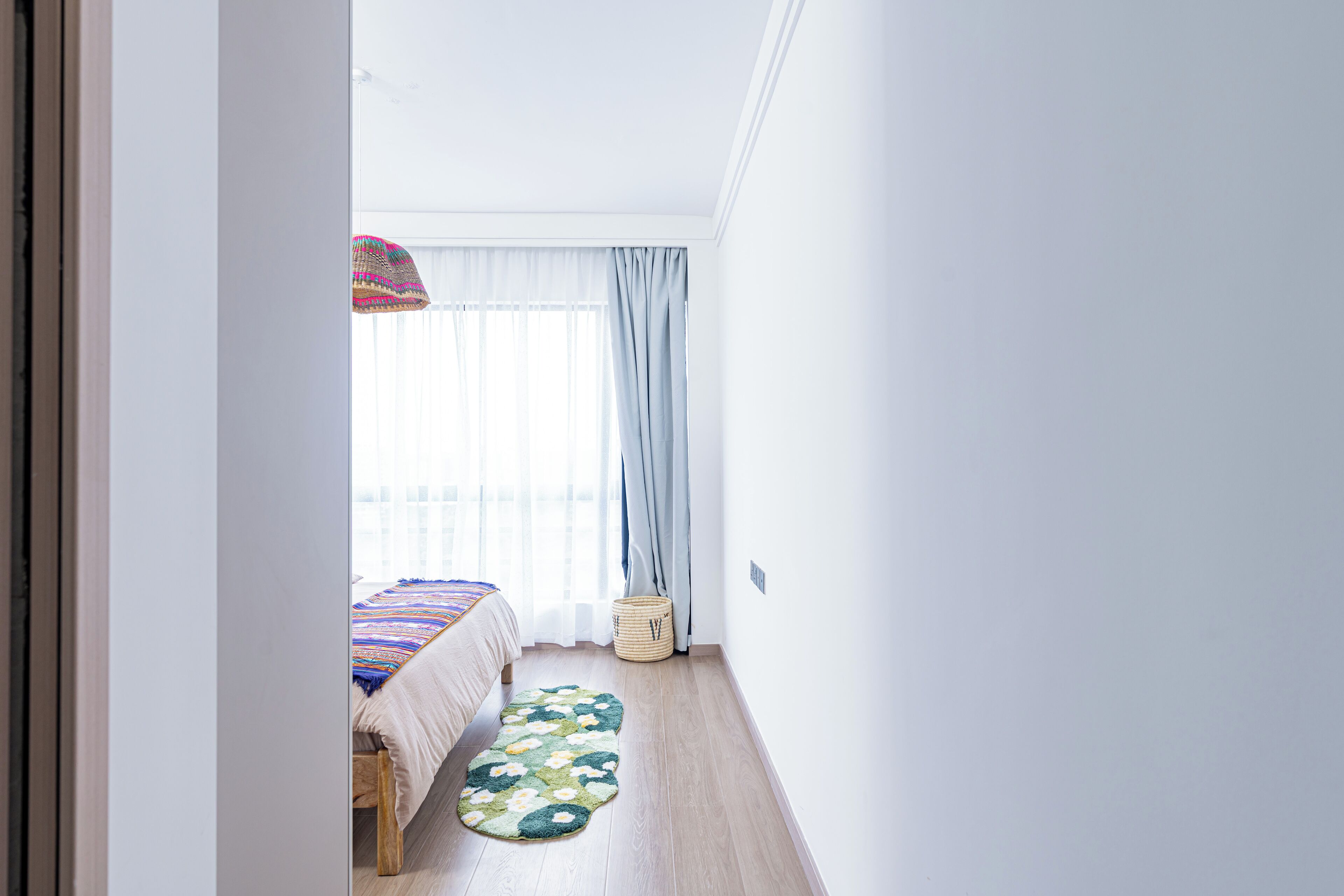Una habitación con cama, ventana con cortinas translúcidas, piso de madera y una pequeña cesta tejida.