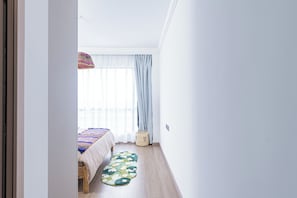 A room with a bed, a window with sheer curtains, a wooden floor, and a small woven basket.