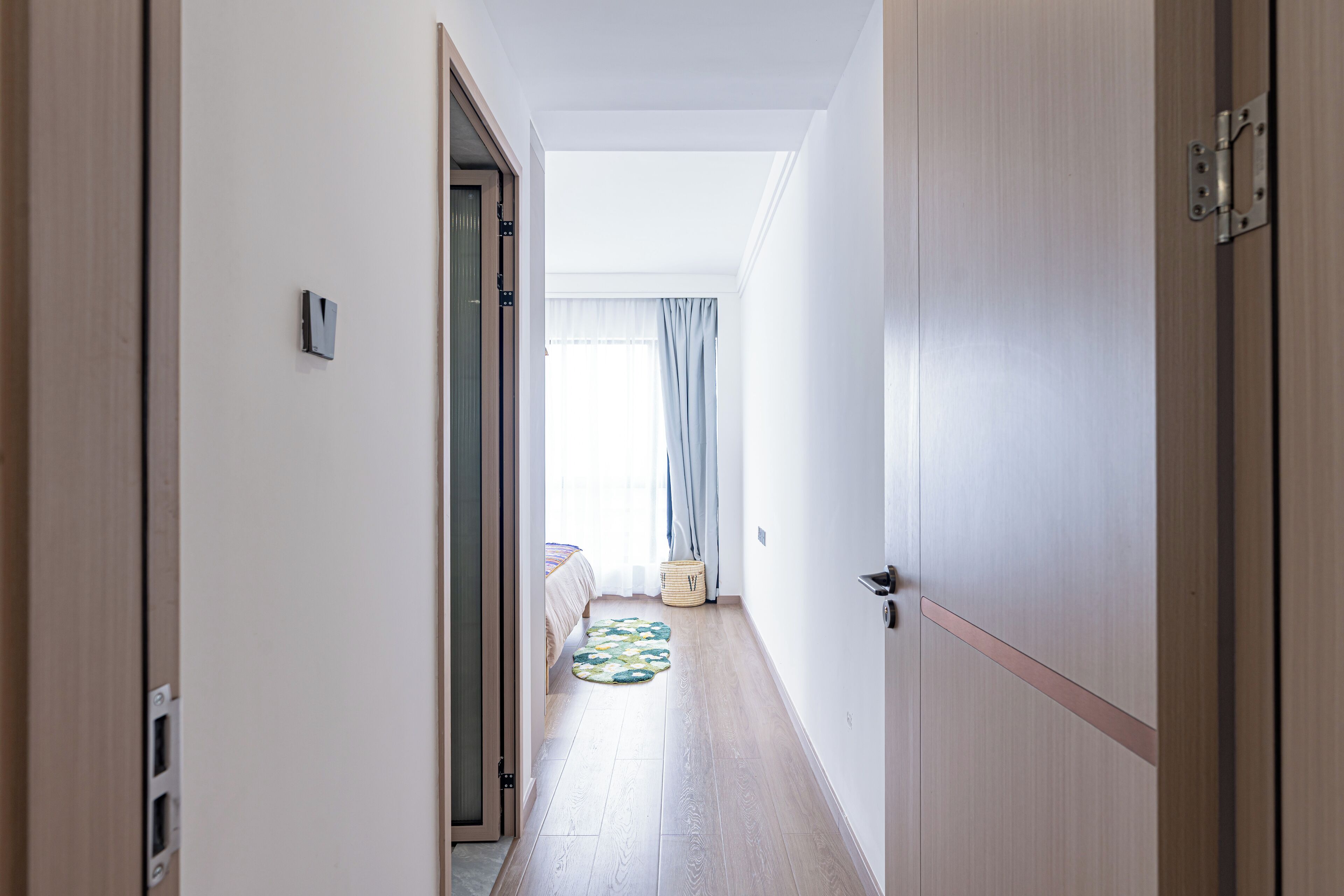A narrow hallway with wooden flooring, a bed with a patterned bedspread, a small basket, and a window with curtains.
