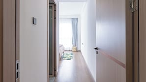 A narrow hallway with wooden flooring, a bed with a patterned bedspread, a small basket, and a window with curtains. - 2 Bedroom - Spacious and Modern - Riverside One (Nairobi)