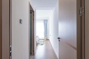 A narrow hallway with wooden flooring, a bed with a patterned bedspread, a small basket, and a window with curtains.