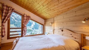 A wooden cabin bedroom with a large bed, plaid curtains, and a window overlooking a snowy mountain.