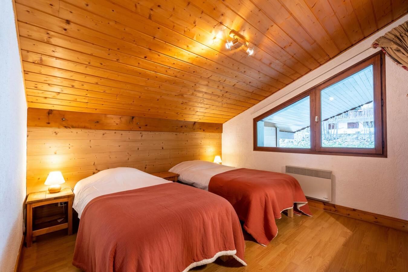 Quarto com teto e paredes de madeira, duas camas com roupa de cama vermelha, uma mesa de cabeceira com abajur e uma janela com vista para um edifício.