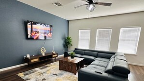 A modern living room with a sectional sofa, a coffee table, a TV mounted on the wall, and a ceiling fan. - Sandy Retreat for family and groups Austin (Round Rock)
