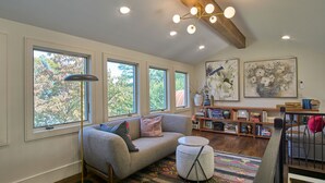 Una sala de estar acogedora con un sofá, una mesita de centro y una estantería llena de libros. Amplias ventanas permiten que entre la luz natural.