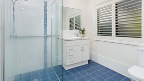 Een badkamer met een douchecabine van glas, een wit wastafeltje met een spiegel erboven en een raam met blinds.