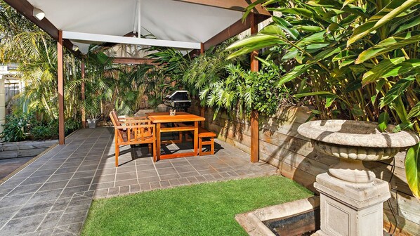 Een terras buiten met een houten tafel en stoelen, omgeven door weelderig groen en een stenen fontein.