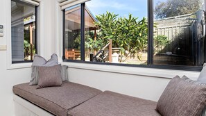 Asiento junto a la ventana con cojines, vista a un jardín y un edificio al fondo.