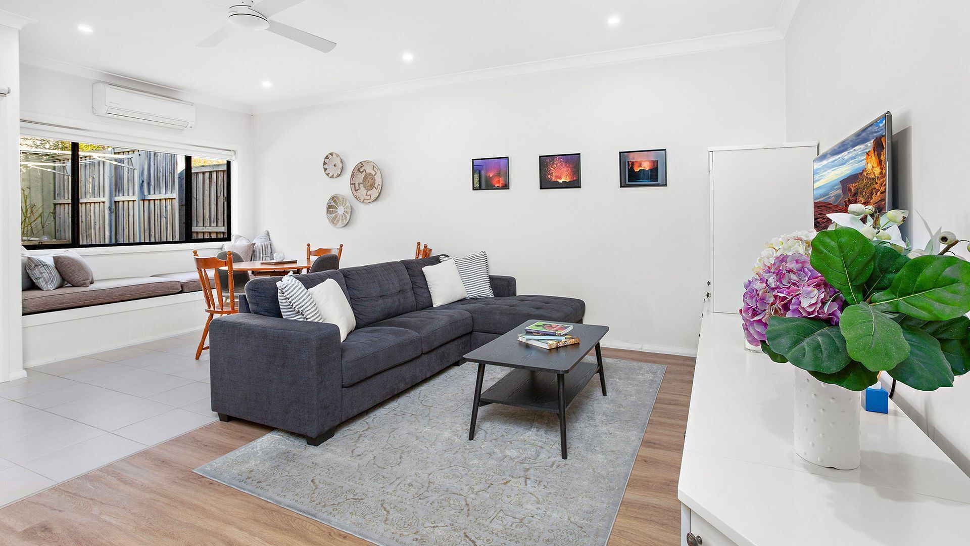 A modern living room with a grey sectional sofa, a coffee table, and a dining area with wooden chairs and a table. There are four framed artworks on the wall and a large window with a view of the outside.