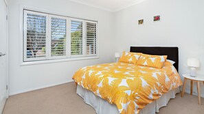 Een slaapkamer met een groot bed, een raam met zonwering, een nachtkastje met een lamp, en twee kleine kunstwerken aan de muur.