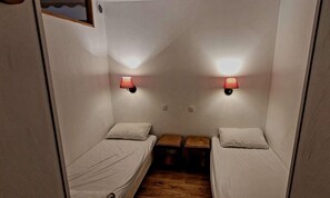 A small, compact room with two single beds, each with a pillow and a nightstand. There are two wall-mounted lamps providing light. The room has a wooden floor and white walls. - Ski-in/ski-out & foot of the slopes, 3rd floor, swimming pool, sauna, balcony, tv, 40m², Les Orres (Les Orres)