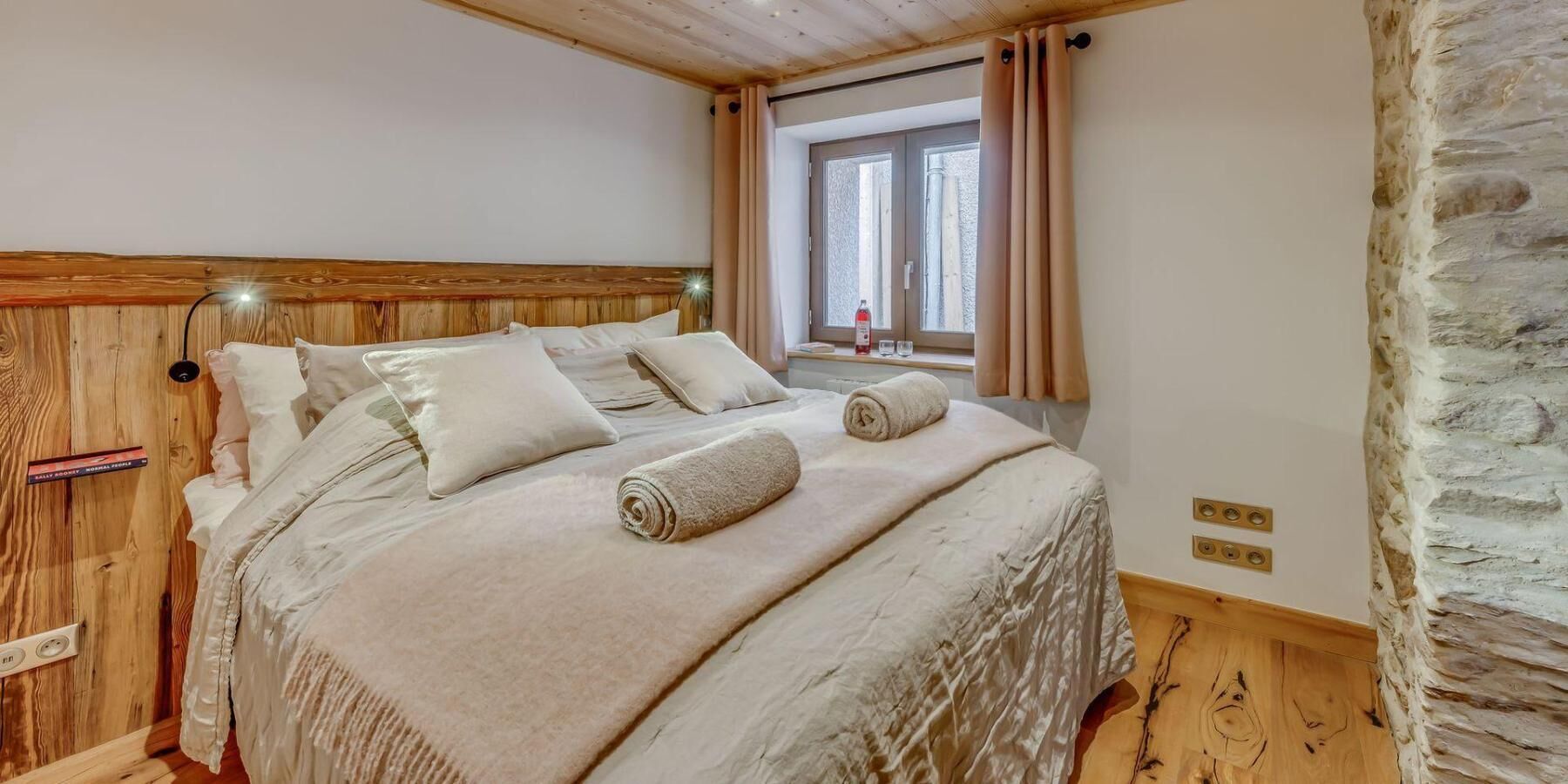 A bedroom with a wooden headboard, a bed with beige linens, two rolled towels, a bedside table with a bottle and glasses, and a window with curtains.