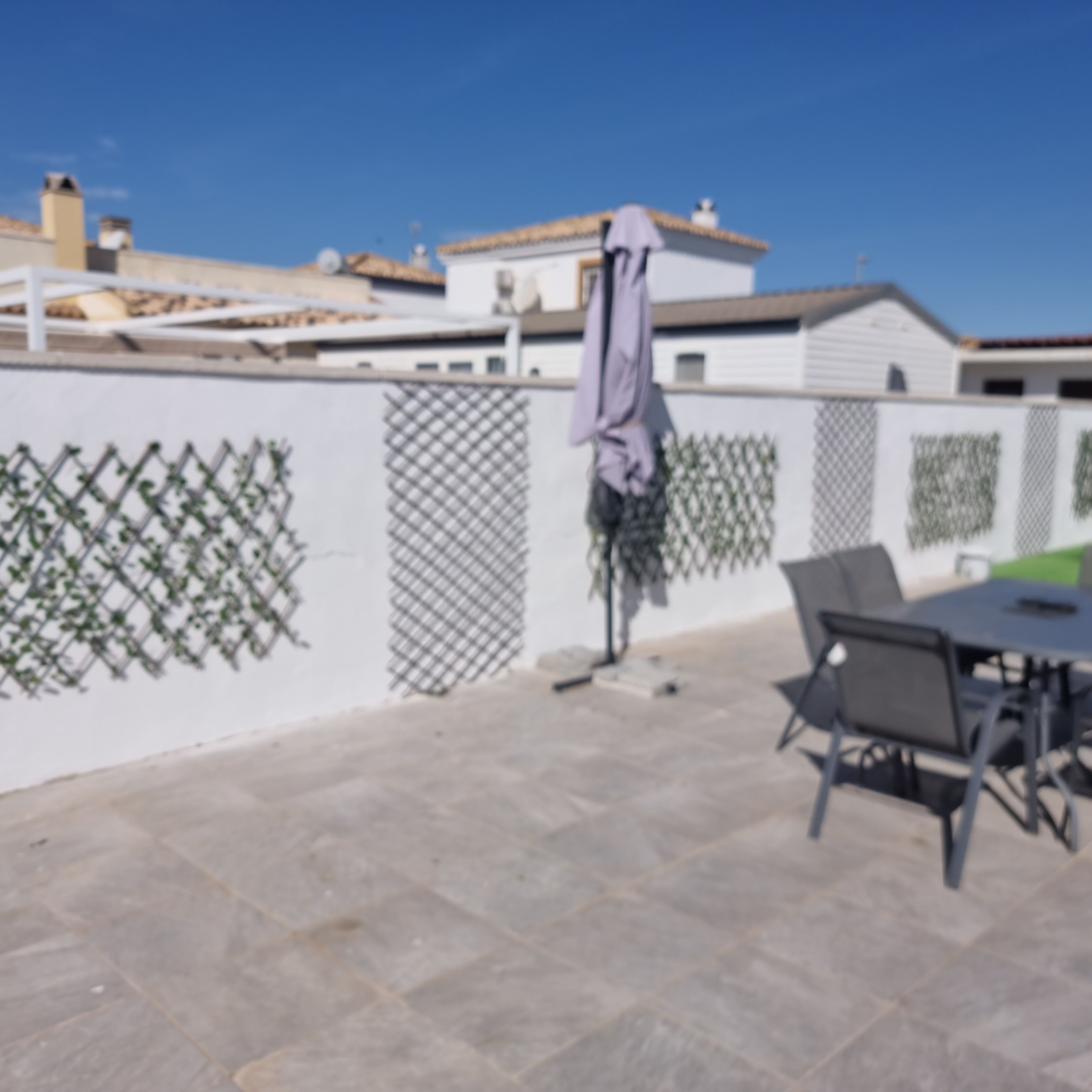 A patio area with a table and chairs, a white fence with greenery, and a building in the background.