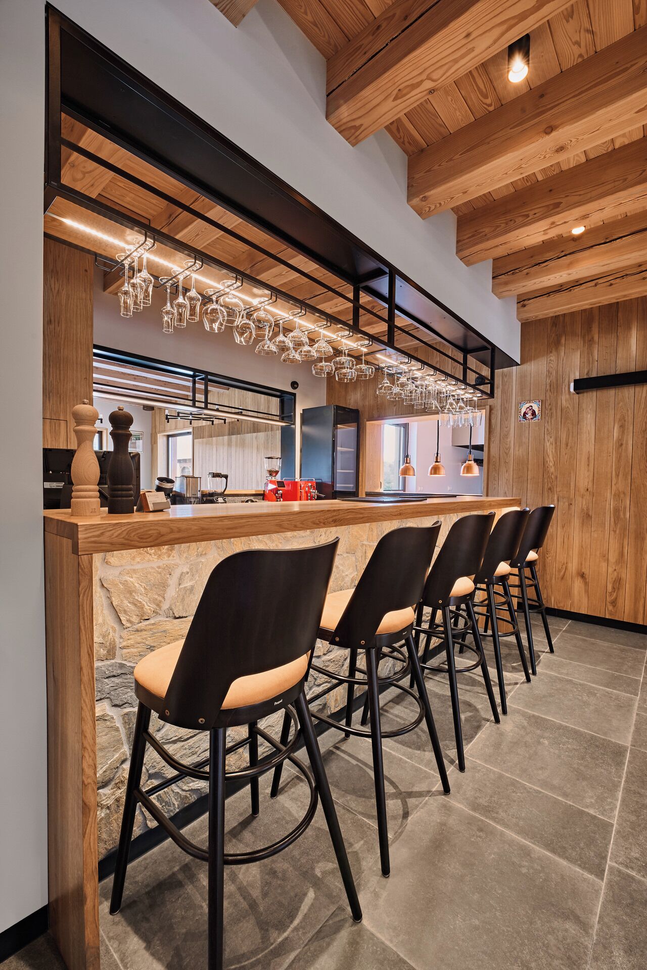 A bar area with a stone counter, black bar stools, wooden ceiling beams, and hanging light fixtures.