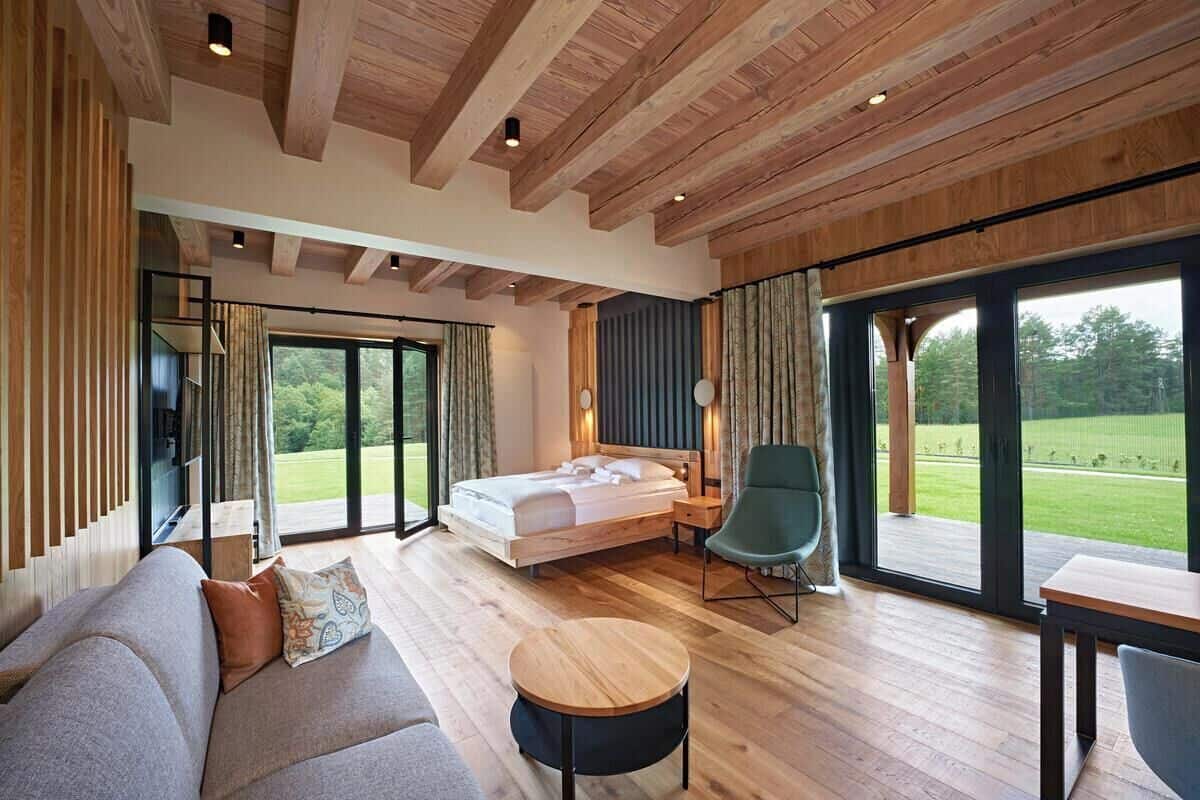 A modern bedroom with a large bed, a sofa, a round table, and a view of a green landscape through large windows.