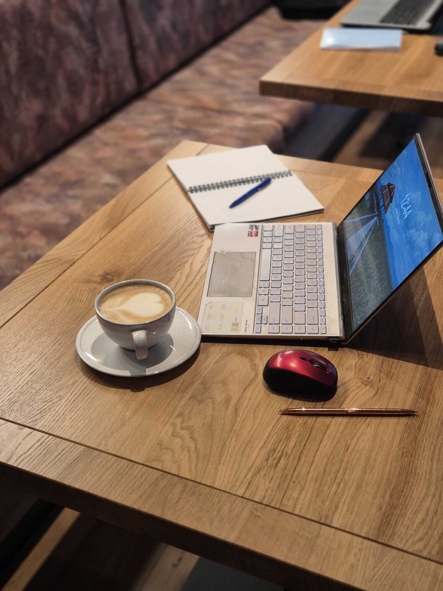 A wooden table with a laptop, a red mouse, a notebook, a pen, and a cup of coffee.