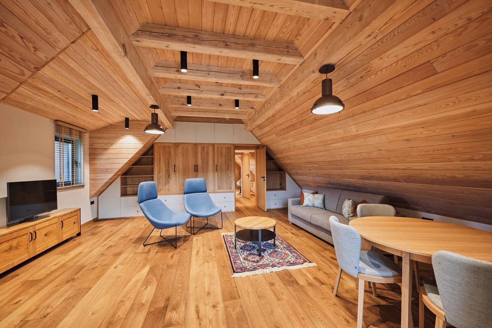 Une pièce mansardée spacieuse avec un plancher en bois, une table à manger, des chaises bleues, un canapé et un téléviseur.