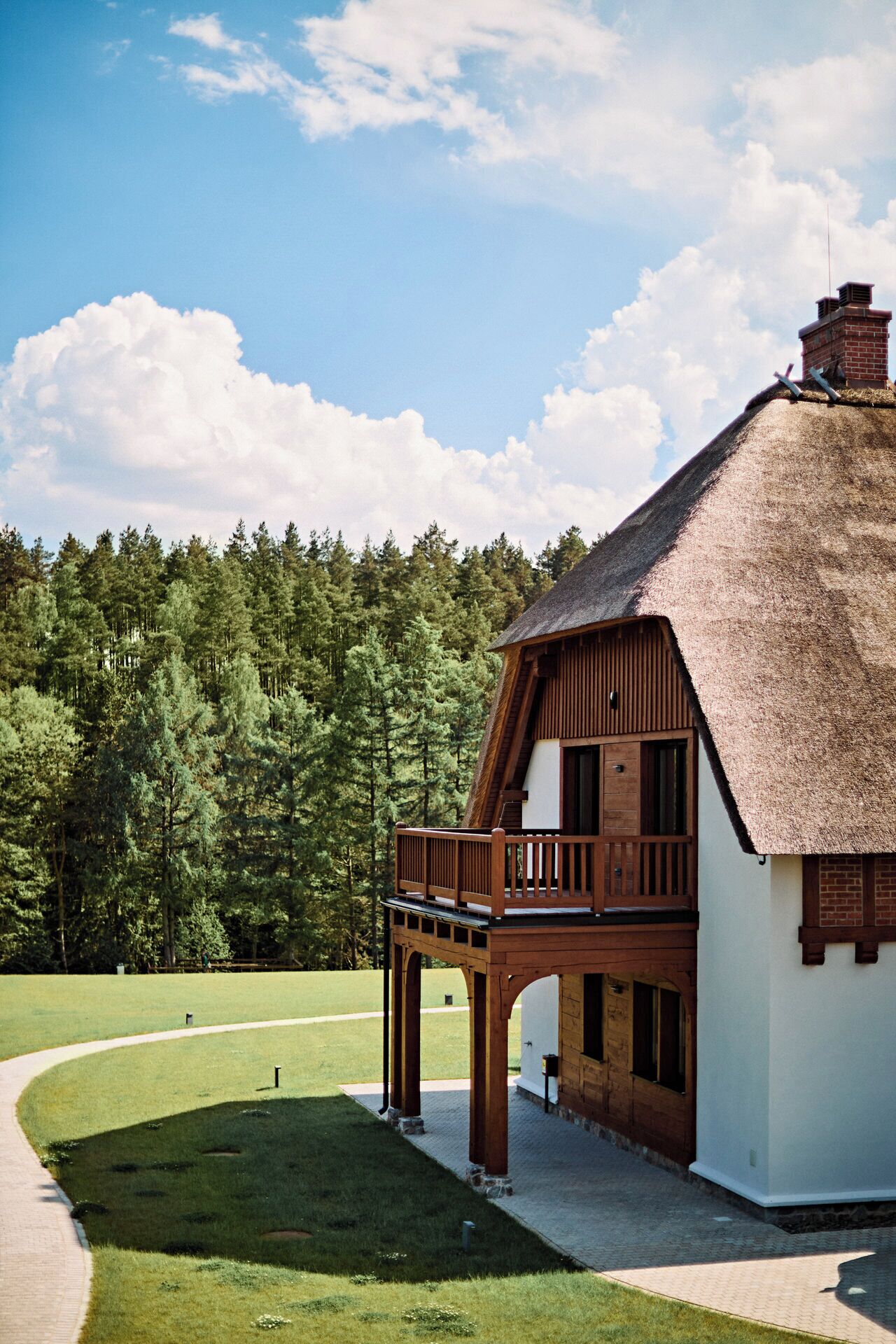 A thatched-roof building with a wooden balcony, surrounded by a grassy area and a forested backdrop.