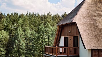 A thatched-roof building with a wooden balcony, surrounded by a grassy area and a forested backdrop.