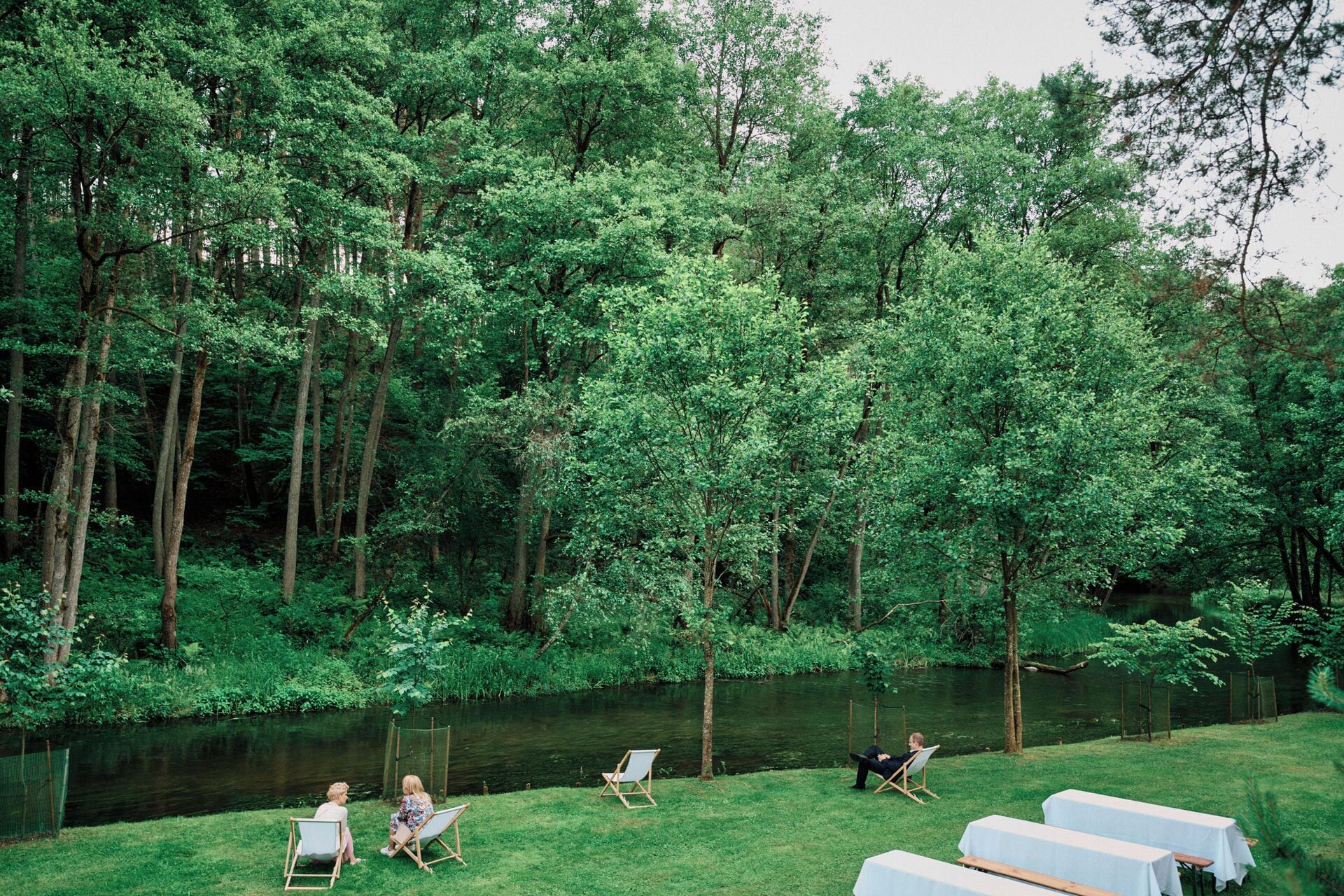 A serene outdoor setting with a grassy area, a pond, and trees. There are two chairs and a table set up for dining.