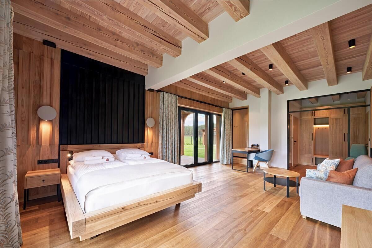A spacious bedroom with a large bed, wooden ceiling beams, and a view of the outdoors through a sliding door.