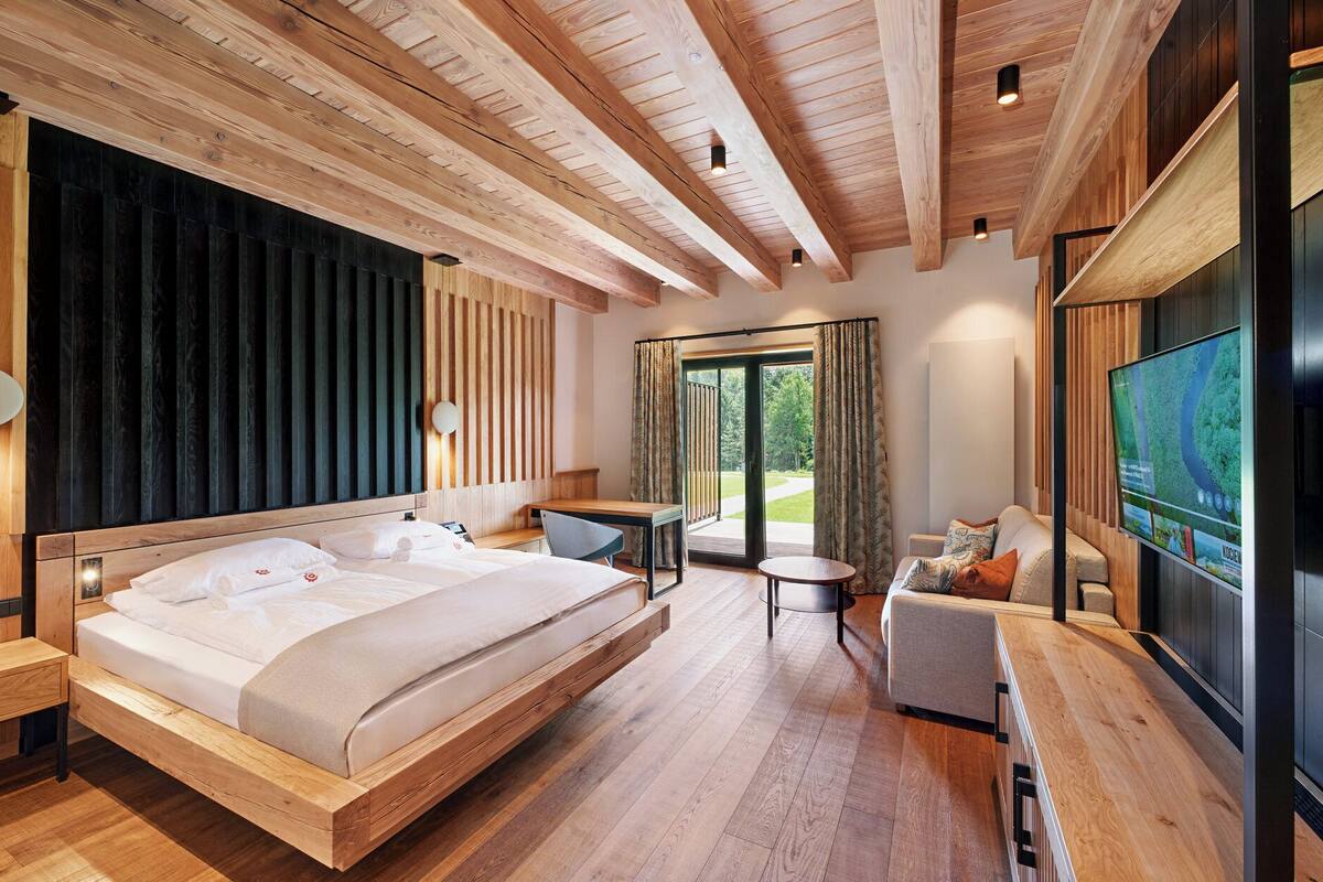 A modern hotel room with a wooden ceiling, a large bed, a desk, a chair, and a TV.