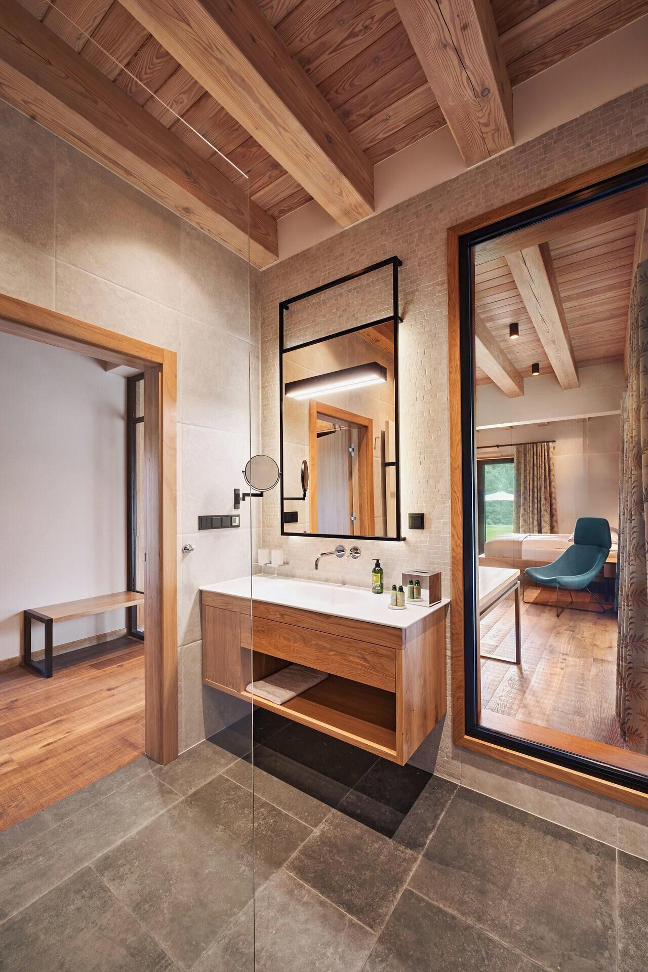Une salle de bain moderne avec un meuble-lavabo en bois, un grand miroir et une vue sur une chambre à coucher où l’on aperçoit un lit et une chaise.