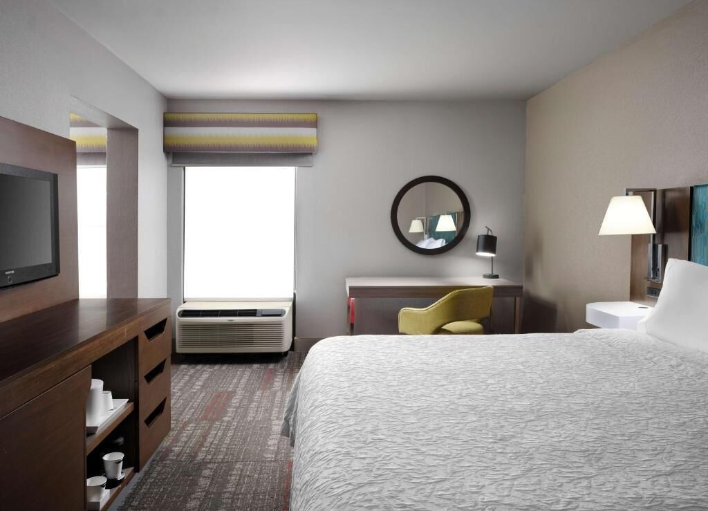 Ein Hotelzimmer mit einem Bett, einem Schreibtisch mit Stuhl, einem Fernseher, einem Spiegel und einem Fenster.