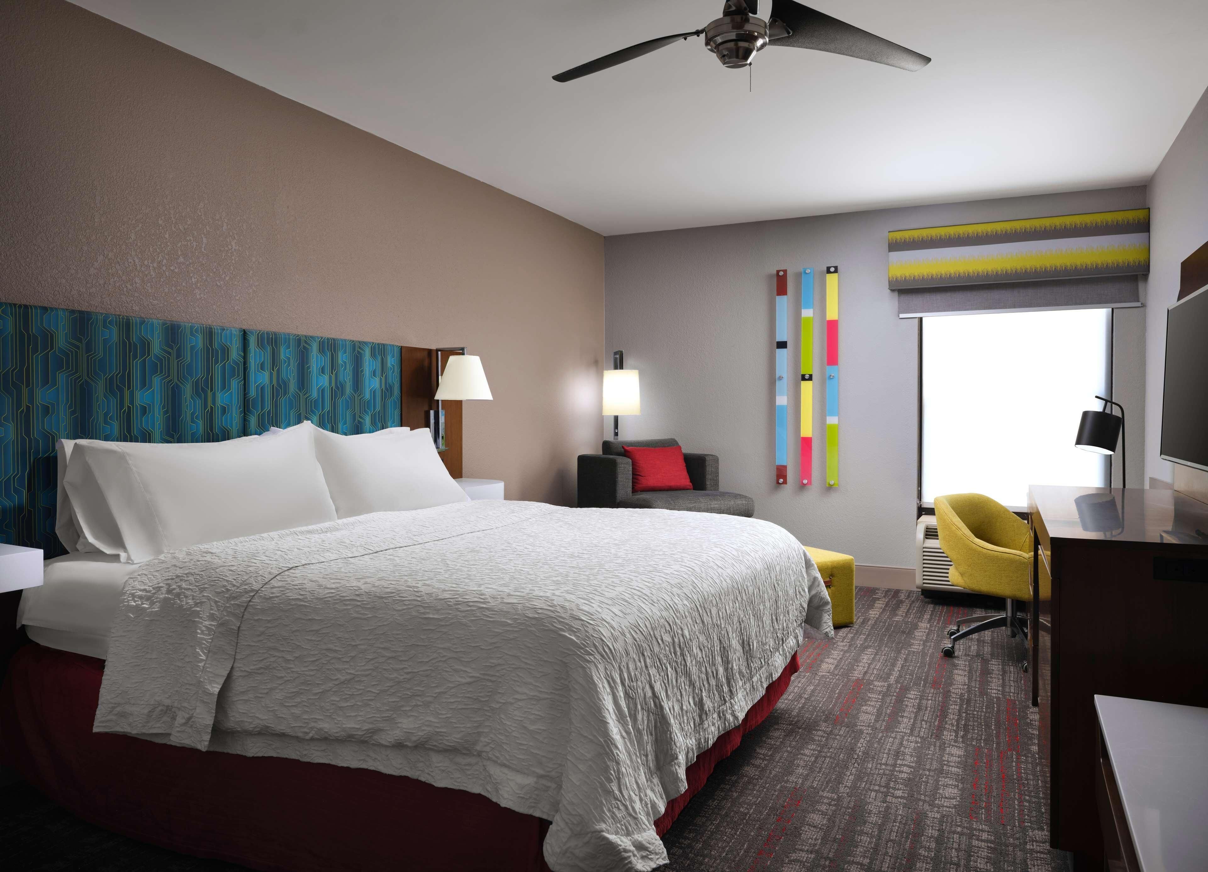 Ein Hotelzimmer mit Bett, Schreibtisch, Stuhl, Deckenventilator, Fernseher und Fenster.
