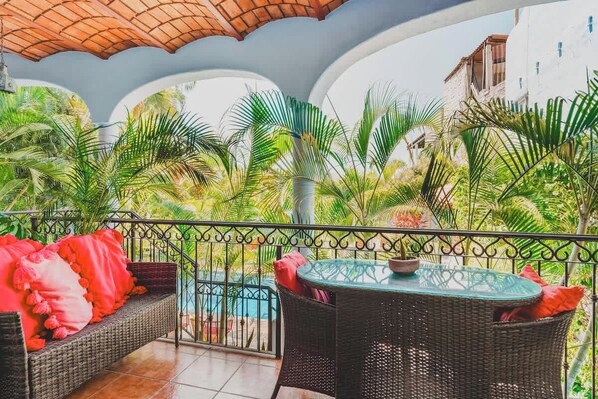 A balcony with wicker furniture, a glass table, and a view of a pool and greenery. - Casa Del Morro - Studio Oasis with heated pool (Sayulita)