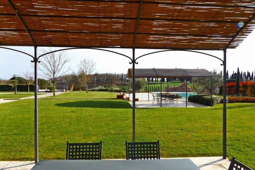 Un espacio de estar techado al aire libre con una mesa y sillas, rodeado por un césped cuidado y una vista distante de un edificio con piscina.