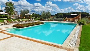 Una piscina rectangular amb un solàrium enrajolat, envoltada de gandules i ombrelles, amb un fons de paisatge de trees i un cel serè.